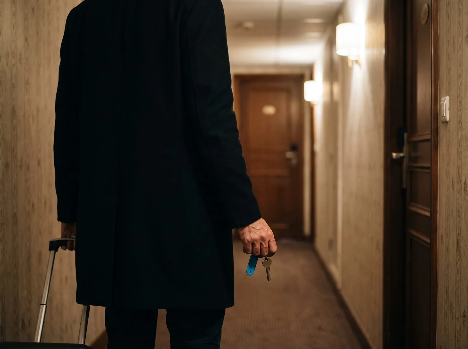 La nuit, un client remonte le couloir d’un hôtel avec sa valise et la clé de sa chambre